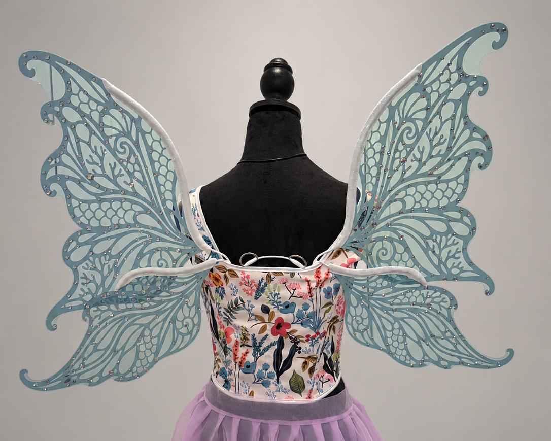 A fairy costume displayed on a black mannequin torso, featuring large light blue lace wings, a floral embroidered bodice, and a lavender tulle skirt against a plain light gray background.