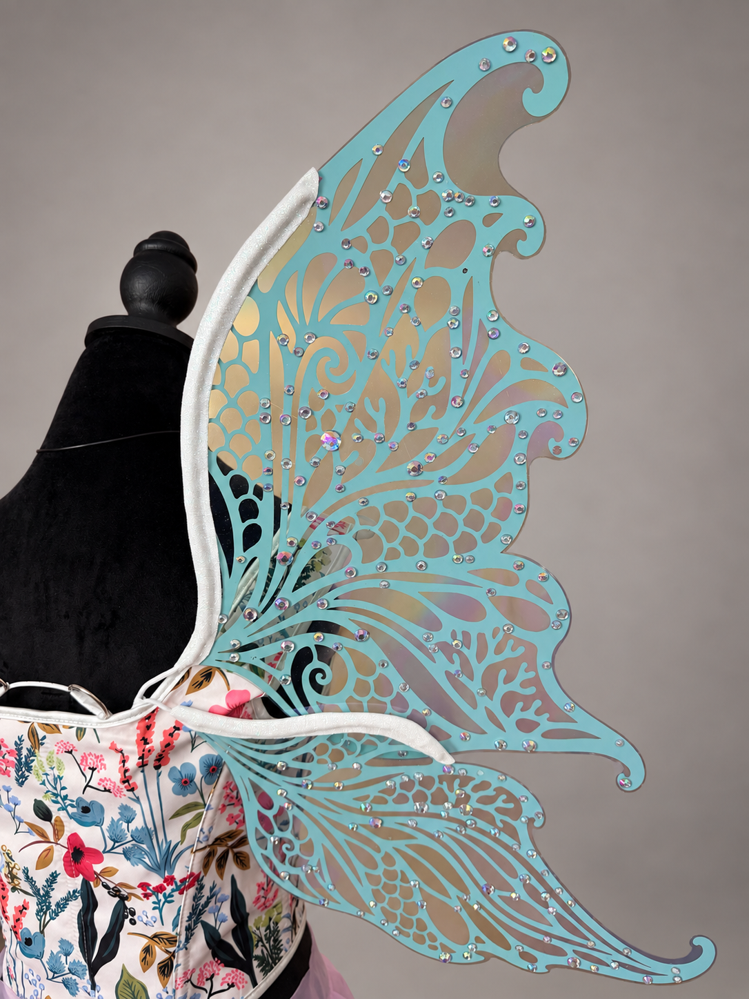 A large decorative wing-like accessory with an intricate filigree pattern, shown attached to a mannequin torso. The main visible color is a light aqua/turquoise tone with small silver rhinestone accents, photographed against a plain taupe-gray background.