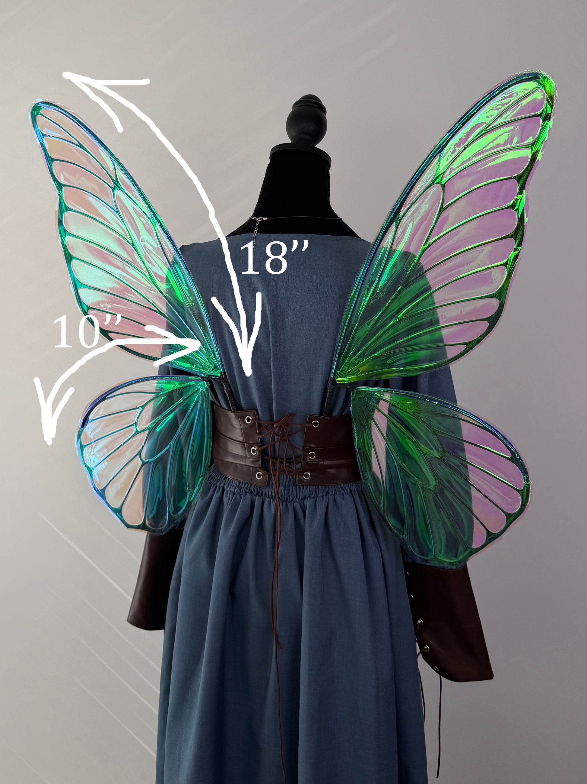 A mannequin wearing a blue dress with a brown lace-up waist and large translucent fairy wings in green, blue, and pink. White arrows and text indicate wing measurements: 18 inches tall and 10 inches wide.