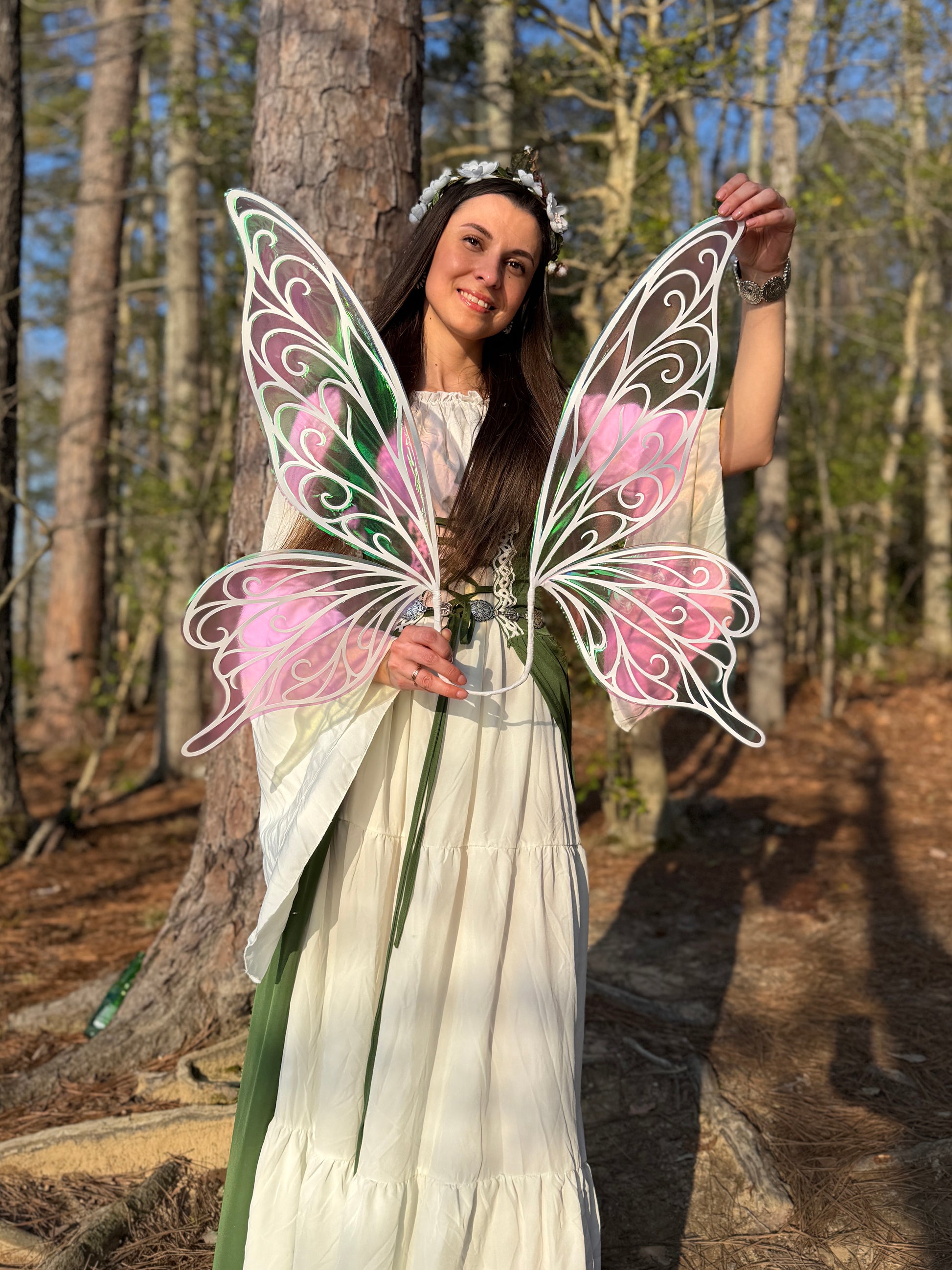 A woman in a white dress and floral headpiece holds up a large fairy wing prop with white filigree outlines and pink and green patterned sections, standing outdoors in a wooded area.