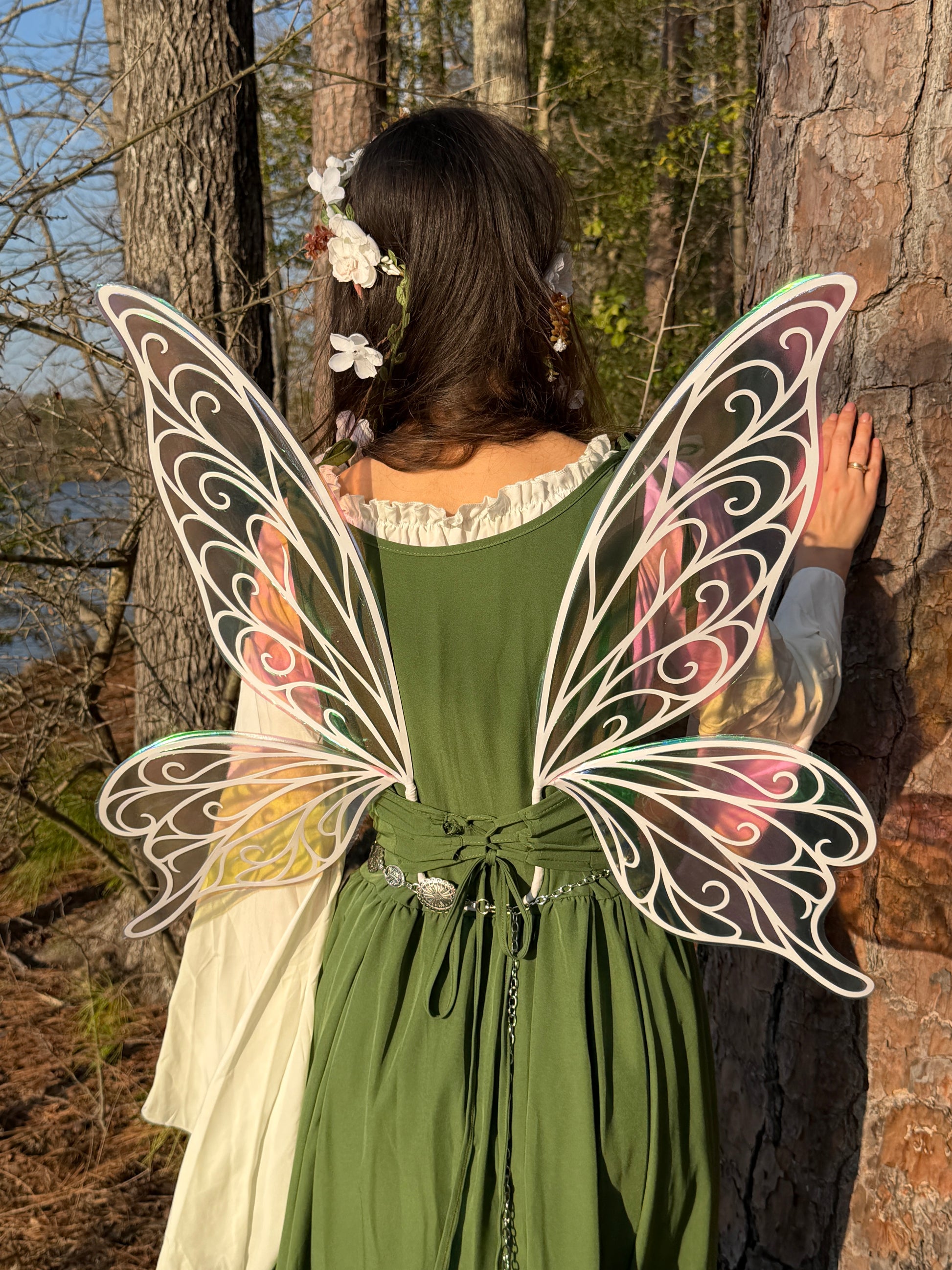 A woman in a forest setting, viewed from behind, wearing a green dress and large decorative fairy wings with white filigree and pastel pink, yellow, and blue accents. She has white flowers in her hair and is leaning against a tree trunk.