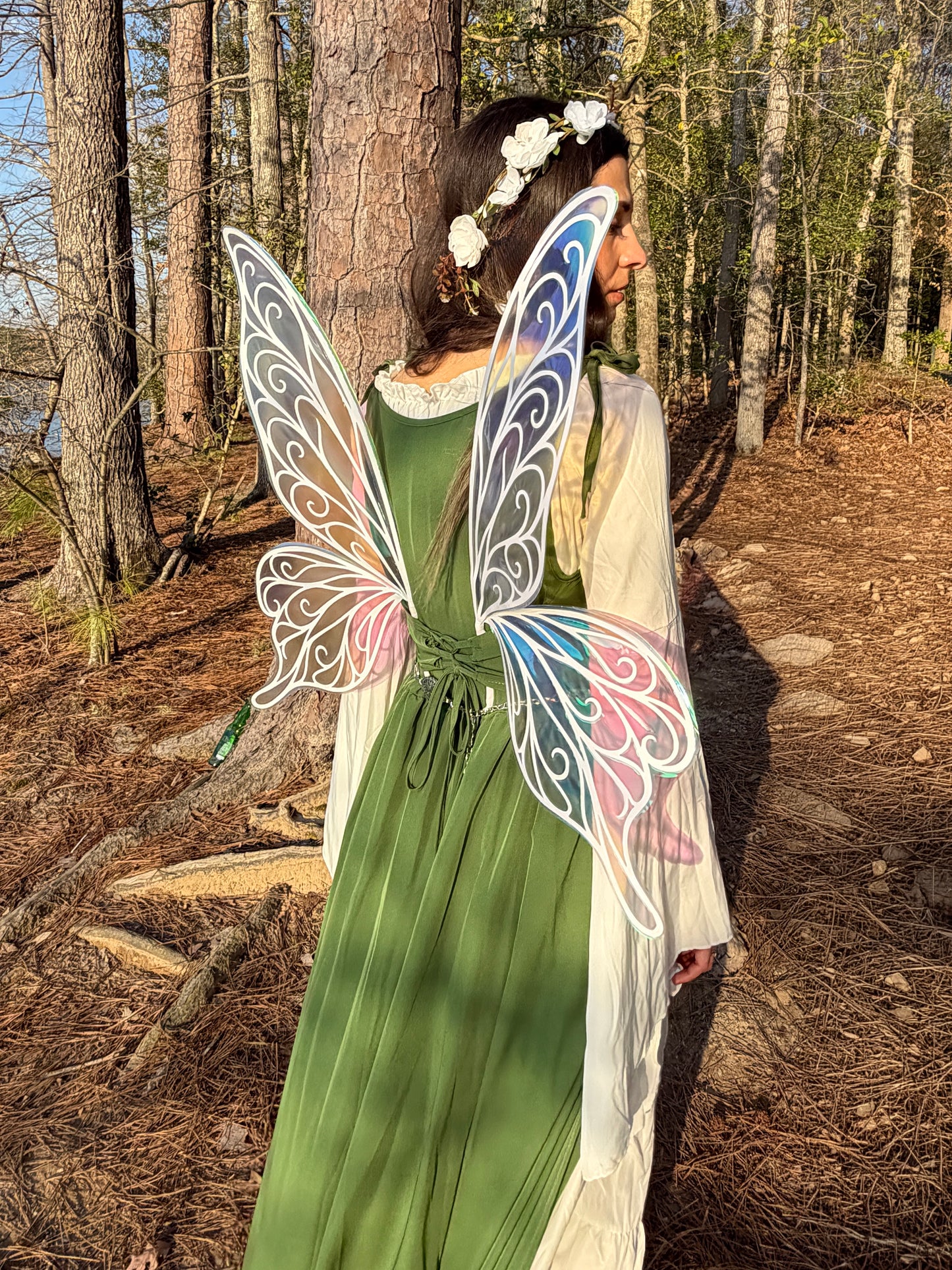 A woman standing in a forest wearing a green dress, white sheer overlay, and large decorative butterfly wings with white outlines and pastel blue, pink, and purple accents.