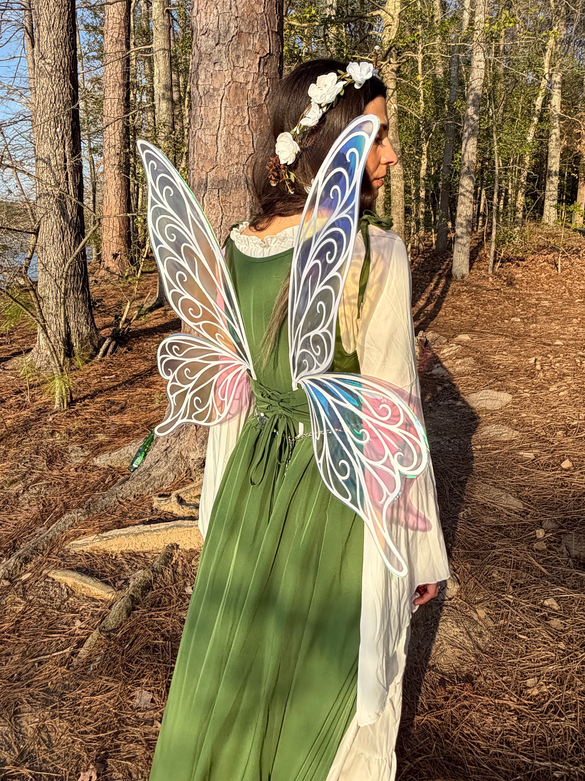 A woman standing in a forest wearing a green dress, white sheer overlay, and large decorative butterfly wings with white outlines and pastel blue, pink, and purple accents.