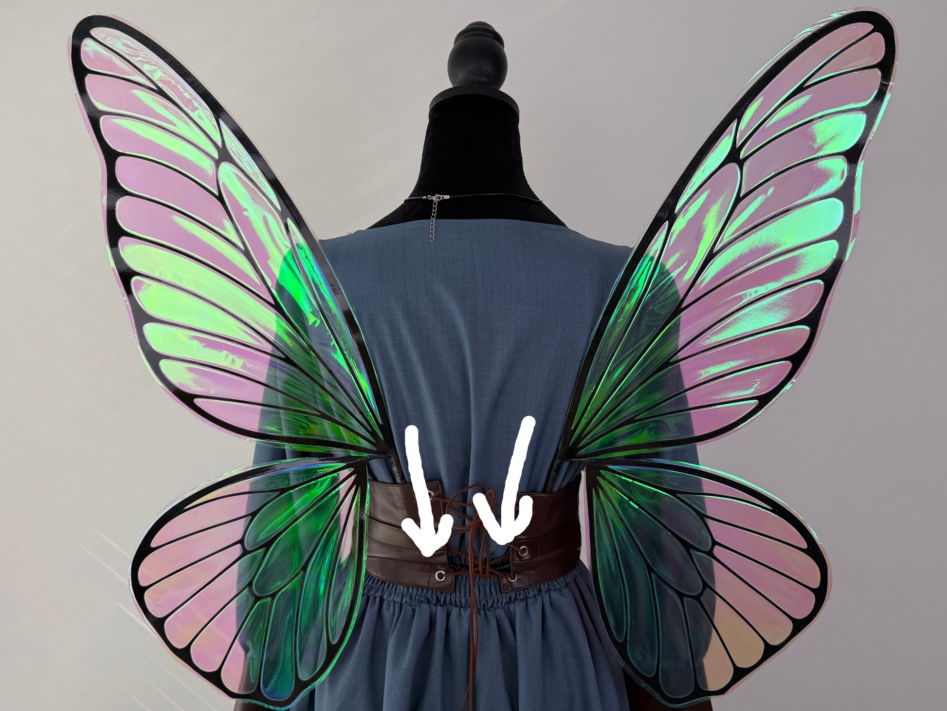 A pair of large fantasy butterfly wings displayed on a mannequin from the back, attached to a blue dress with a brown belt. White arrows point to the wing attachments.